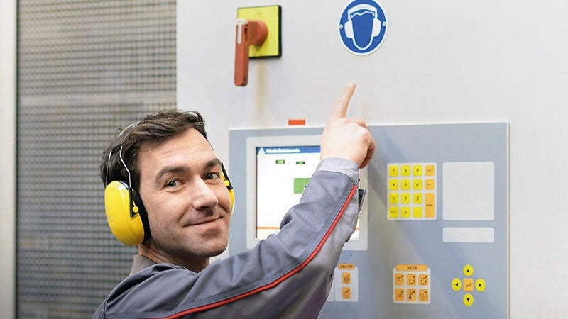 ein Mann mit Gehörschutz steht in einem Maschinenraum und deutet auf das Symbol für das Tragen von Gehörschutz