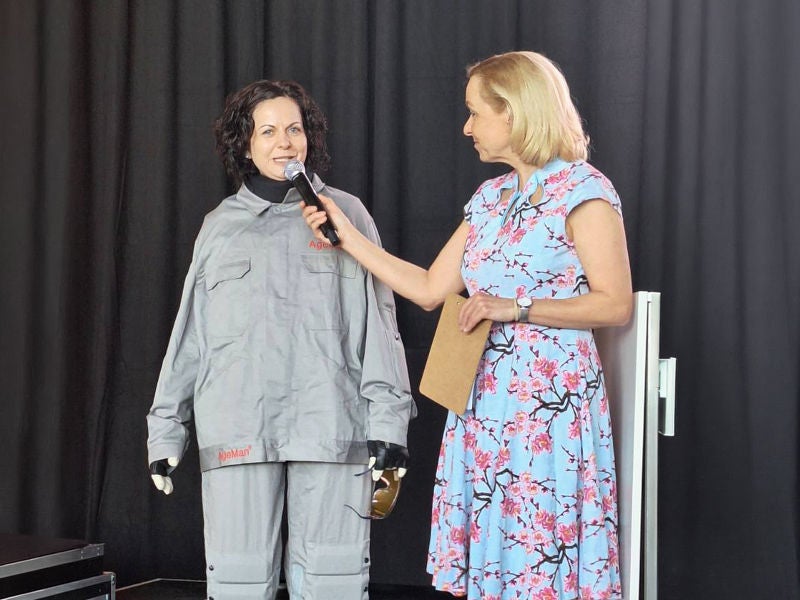Eine Person in einem blauen Kleid mit Blumenmuster hält ein Mikrofon vor eine andere Person, die einen grauen Schutzanzug trägt und auf einer Bühne steht.