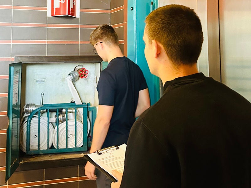 Zwei Personen mit kurzen Haaren und dunkler Kleidung inspizieren einen geöffneten Wandhydrantenkasten mit aufgerollten Schläuchen in einem Flur mit braun-grauen Kacheln.