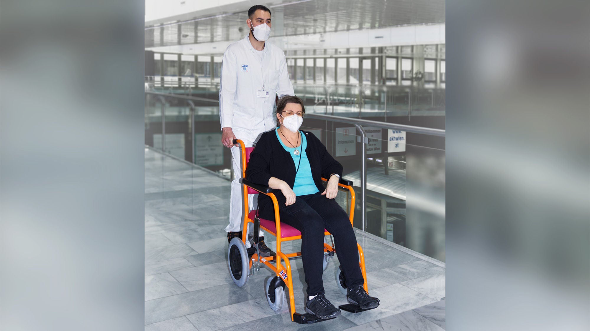 Ein Pfleger mit Mundschutz schiebt den Rollstuhl mit einer darinsitzenden Frau, die ebenfalls Mundschutz trägt. Das herumliegende Ambiente sieht nach einer Klinik aus.