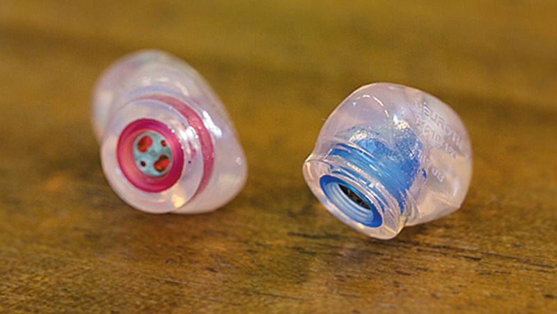 Zwei individuell angepasste Gehörschutz-Otoplastiken in Rot und Blau auf einem Holzuntergrund