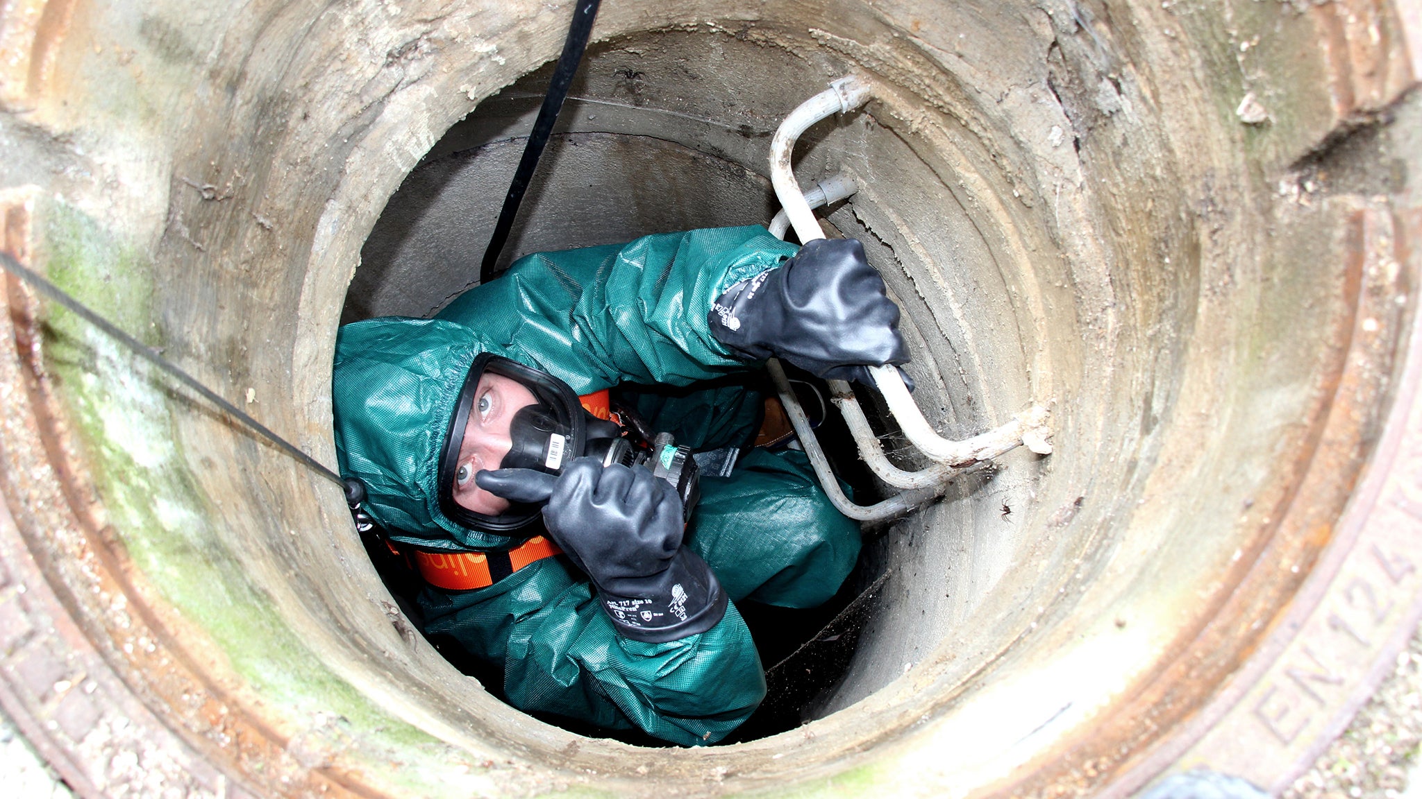 Mann mit Schutzanzug, Schutzbrille und Schutzhandschuhen klettert in Kanal hinab