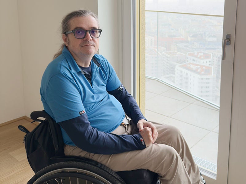 Eine Person mit Brille und langem Haar sitzt in einem Rollstuhl neben einer großen Glasfront mit Blick auf eine Stadtlandschaft.