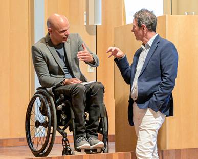 Zwei Personen führen in einem modernen Innenraum ein Gespräch, wobei eine Person im Rollstuhl sitzt und die andere Person mit einer Hand in der Hosentasche zuhört.