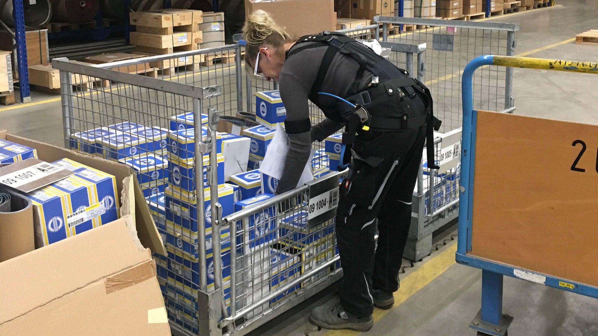 Eine Arbeiterin mit angelegtem Exoskelett schlichtet Sixpacks Bier in eine Gitterboxpalette 