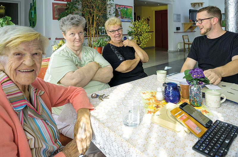 mehrere Seniorinnen sitzen an einem Tisch zusammen, daneben die Sicherheitsfachkraft