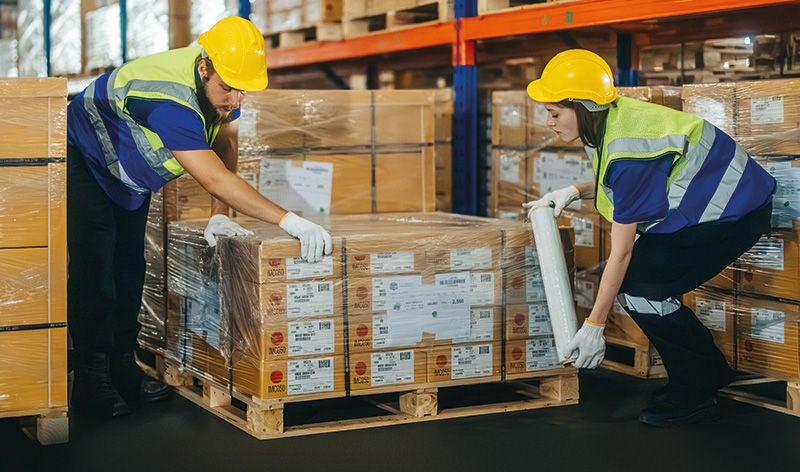 Ein Arbeiter und eine Arbeiterin packen einen Stapel Kartons mit Plastikfolie ein
