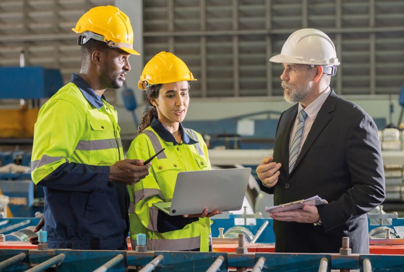 Drei Personen mit Schutzhelmen und Sicherheitskleidung besprechen sich in einer Industriehalle; eine Frau hält einen Laptop, während ein Mann in Anzug und Helm Notizen macht.