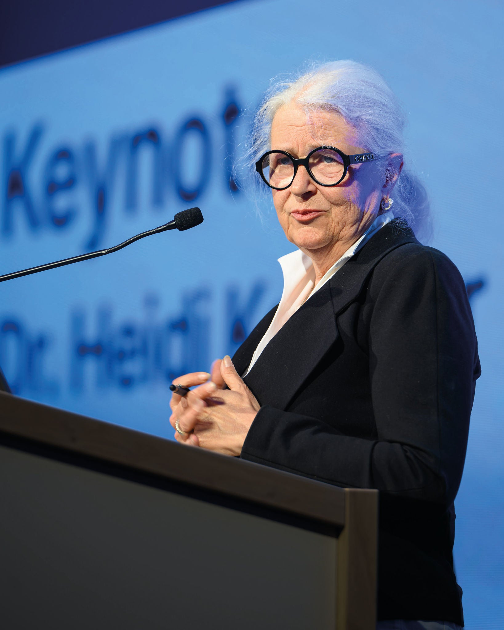 Dr.in Heidi Kastner spricht am Rednerpult vor blauem Hintergrund mit groß eingeblendetem Schriftzug; sie trägt eine schwarze Brille und einen dunklen Blazer.