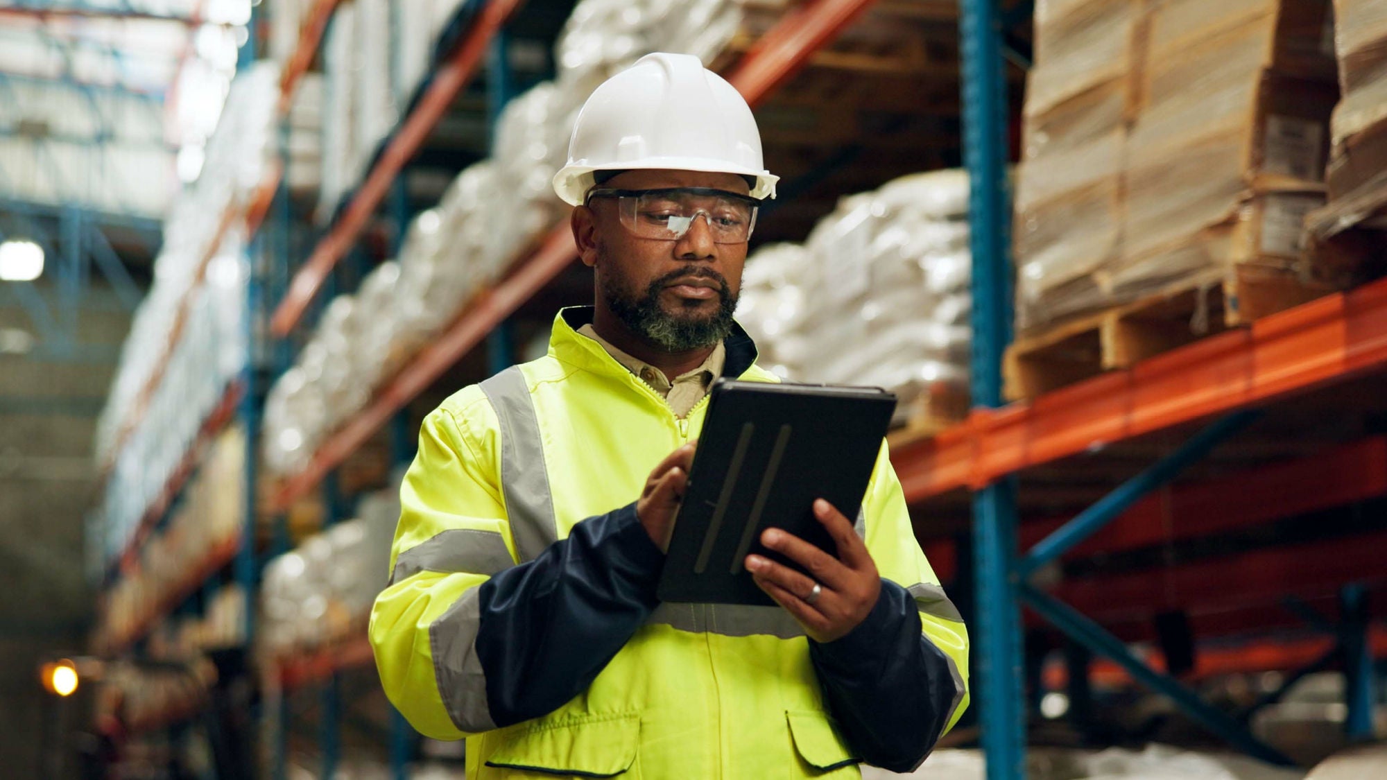 Eine Person in Schutzhelm und Warnweste arbeitet mit einem Tablet in einem Lagerhaus mit Regalen voller verpackter Waren.