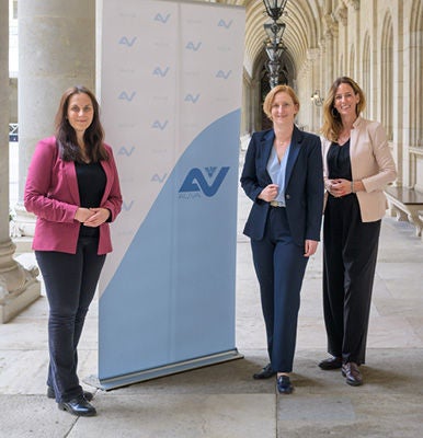 Drei Frauen in Business-Kleidung stehen lächelnd nebeneinander vor einer AUVA-Stellwand.