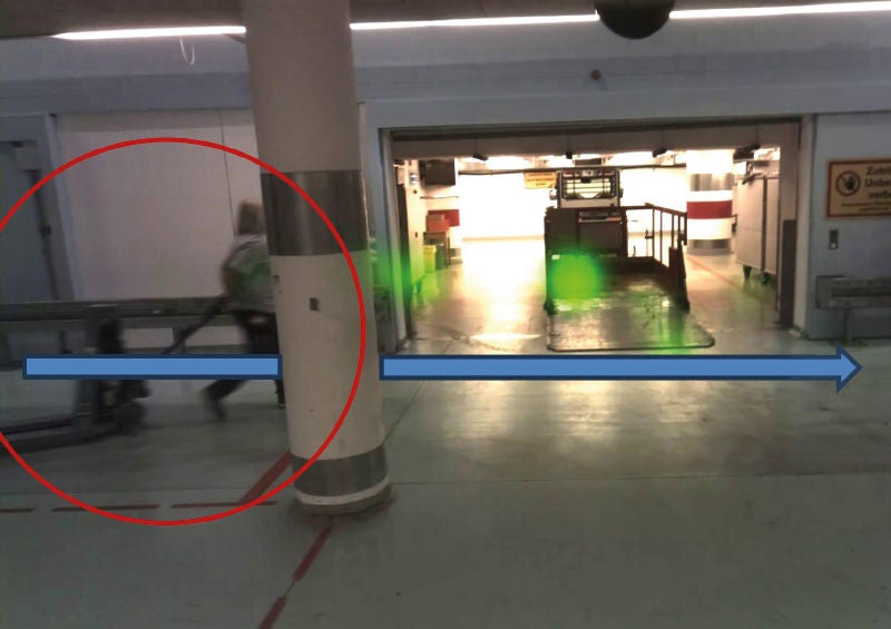 Eine Überwachungsaufnahme zeigt eine Person, die einen Wagen durch einen Raum zieht. Im Hintergrund befindet sich ein gut beleuchteter Bereich mit einem Fahrzeug. Auf dem Bild sind ein roter Kreis und ein blauer Pfeil hinzugefügt, um die Bewegung der Person und den möglichen Fahrweg des Wagens zu markieren.