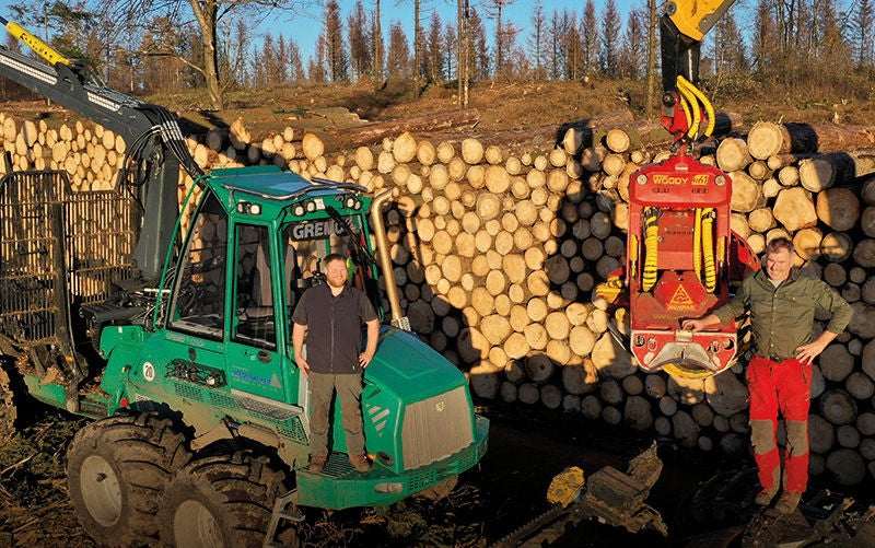 zwei Männer stehen vor einem Stapel Holz und großen Forstmaschinen