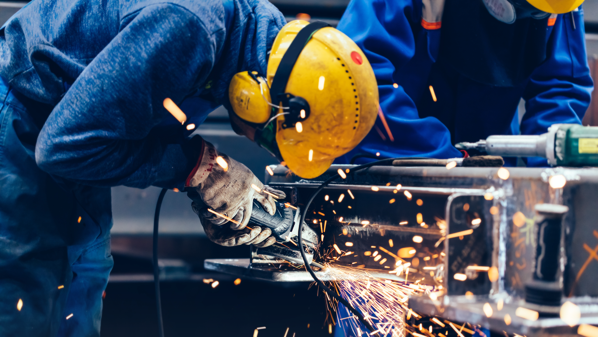 Ein Arbeiter mit Schutzkleidung bestehend aus Helm, Gehörschutz und Handschuhe  arbeitet mit einer Flex. Man sieht Funken sprühen.