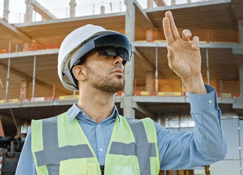 ein Arbeiter mit VR Brille zeig tmit zwei Fingern in die Luft, dahinter sieht man eine Baustelle