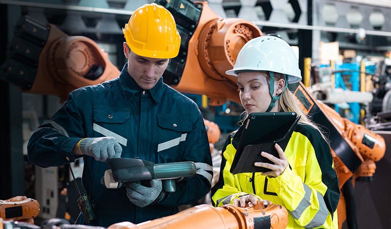 Zwei Beschäftigte mit Schutzhelmen arbeiten gemeinsam mit einem Industrieroboter – symbolisiert Mensch-Maschine-Kollaboration