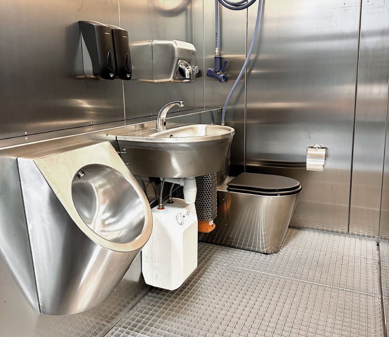 Innenansicht einer mobilen Luxus-Toilette aus Edelstahl mit Urinal, Waschbecken, WC, Desinfektions- und Papierhandtuchspender sowie Duschvorrichtung.