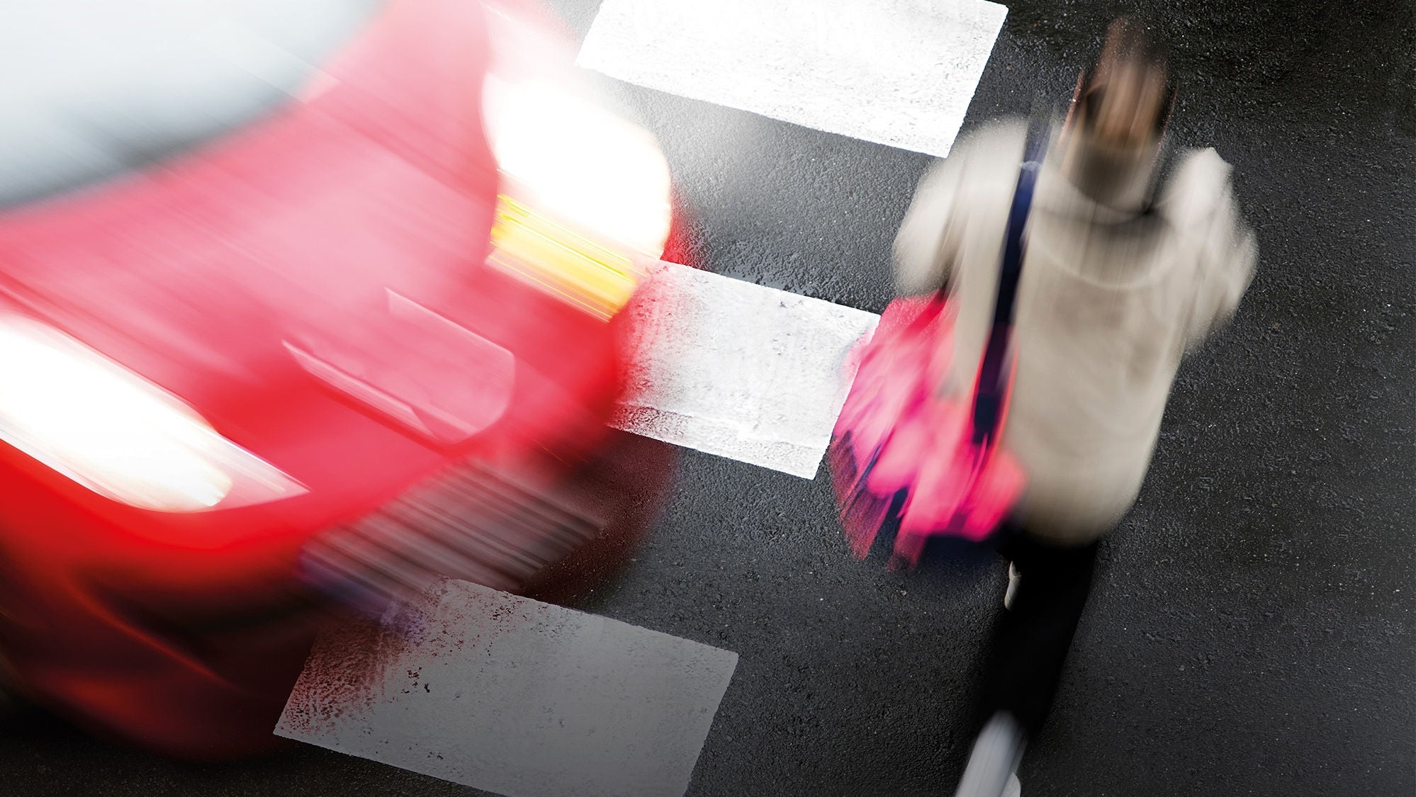 Sicht von oben auf eine Frau, die einen Zebrastreifen überquert, von links nähert sich ein Fahrzeug, beides wirkt verschwommen wie in Bewegung.