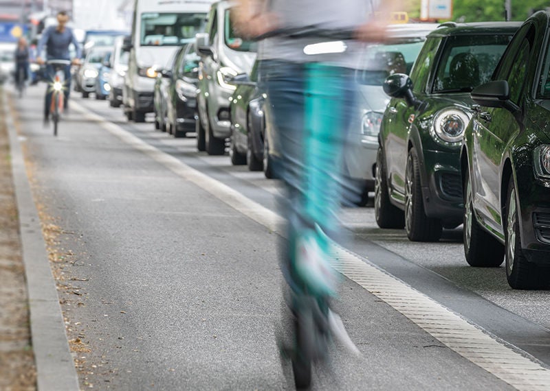 Straße mit vielen Autos, daneben überholen Fahrradfahrer und Scooter