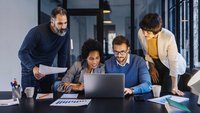 Vier Personen sitzen und stehen an einem Tisch in einem modernen Büro und schauen gemeinsam auf einen Laptop. Eine Frau zeigt mit dem Finger auf den Bildschirm, während ein Mann neben ihr aufmerksam schaut. Eine weitere Frau und ein Mann beugen sich interessiert über die Szene. Auf dem Tisch liegen Papiere, ein Stift und eine Kaffeetasse.