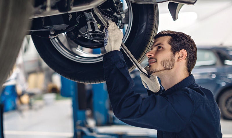 Ein Mechaniker in blauer Arbeitskleidung arbeitet unter einem angehobenen Auto in einer Werkstatt. Er trägt Handschuhe und benutzt einen Schraubenschlüssel, um an der Radaufhängung zu arbeiten.