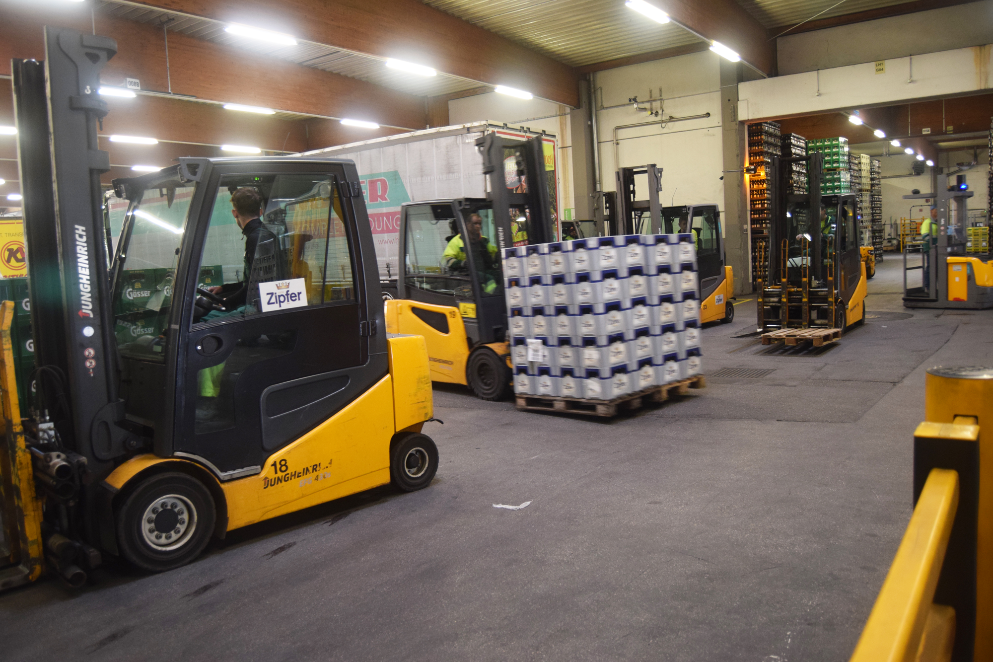 Gabelstabler, einer davon beladen mit Bierkisten, fahren in einer Halle herum, man sieht einen LKW und