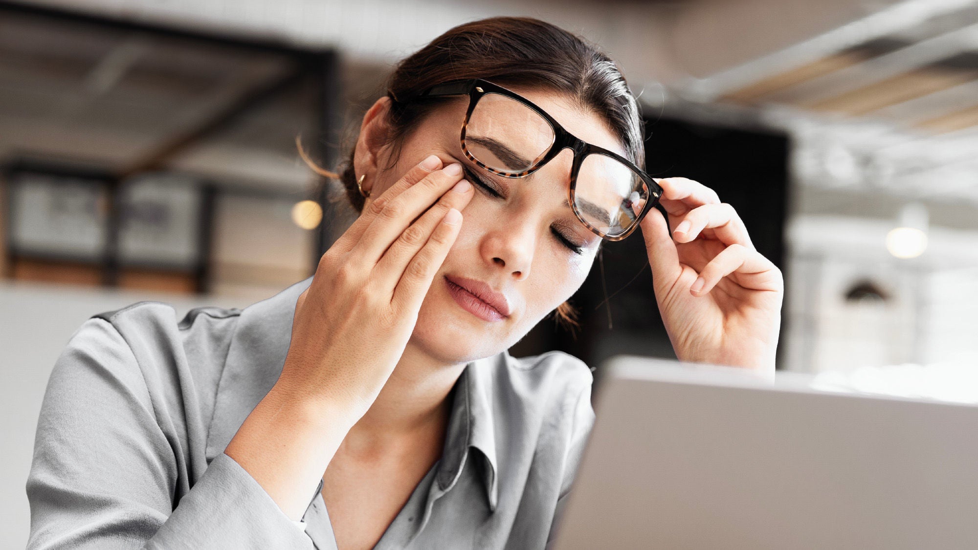 Eine Frau mit Brille reibt sich müde die Augen und sitzt vor einem Laptop, was auf Erschöpfung oder Überanstrengung bei der Bildschirmarbeit hinweist.