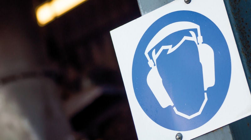 Hinweisschild mit einem stilisierten Kopf, der einen Gehörschutz trägt, kennzeichnet einen lärmintensiven Arbeitsplatz.