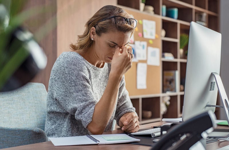 Frau sitzt an einem Schreibtisch vor einem Computer, hält sich die Stirn und wirkt gestresst oder nachdenklich.