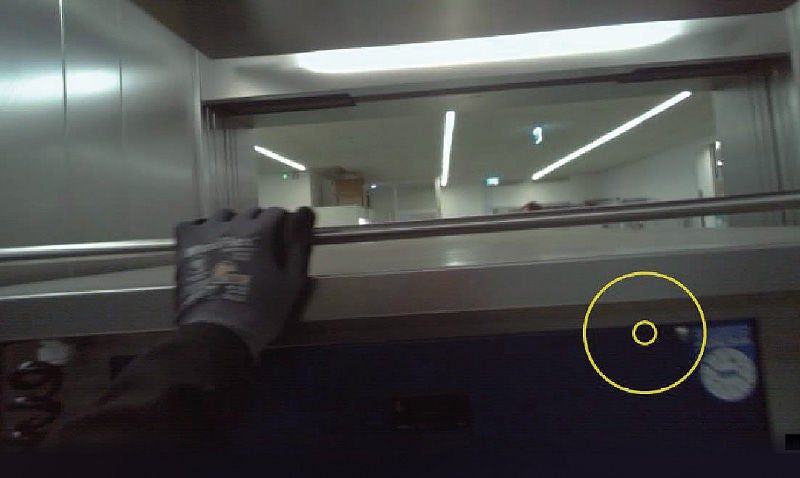 Eine Nahaufnahme einer Person, die mit behandschuhten Händen eine Metallstange greift. Im Hintergrund ist ein industrieller Bereich mit hellen Deckenleuchten zu sehen.