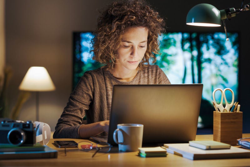 Eine Frau mit lockigem Haar arbeitet abends an einem Laptop, umgeben von Büromaterialien, mit einer Schreibtischlampe als Lichtquelle.