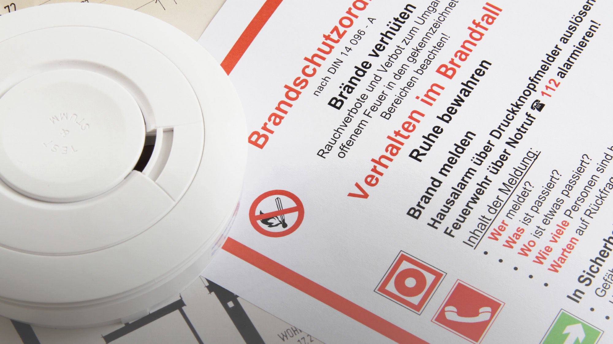 Ein Rauchmelder liegt neben einem Brandschutzordner, der Anweisungen zum Verhalten im Brandfall enthält. Der Text auf dem Dokument betont Ruhe zu bewahren, Brände zu melden und Sicherheitsvorkehrungen zu treffen. Symbole für Notruf, Feuerlöscher und Fluchtwege sind ebenfalls auf dem Papier zu sehen, zusammen mit einem Gebäudegrundriss im Hintergrund.