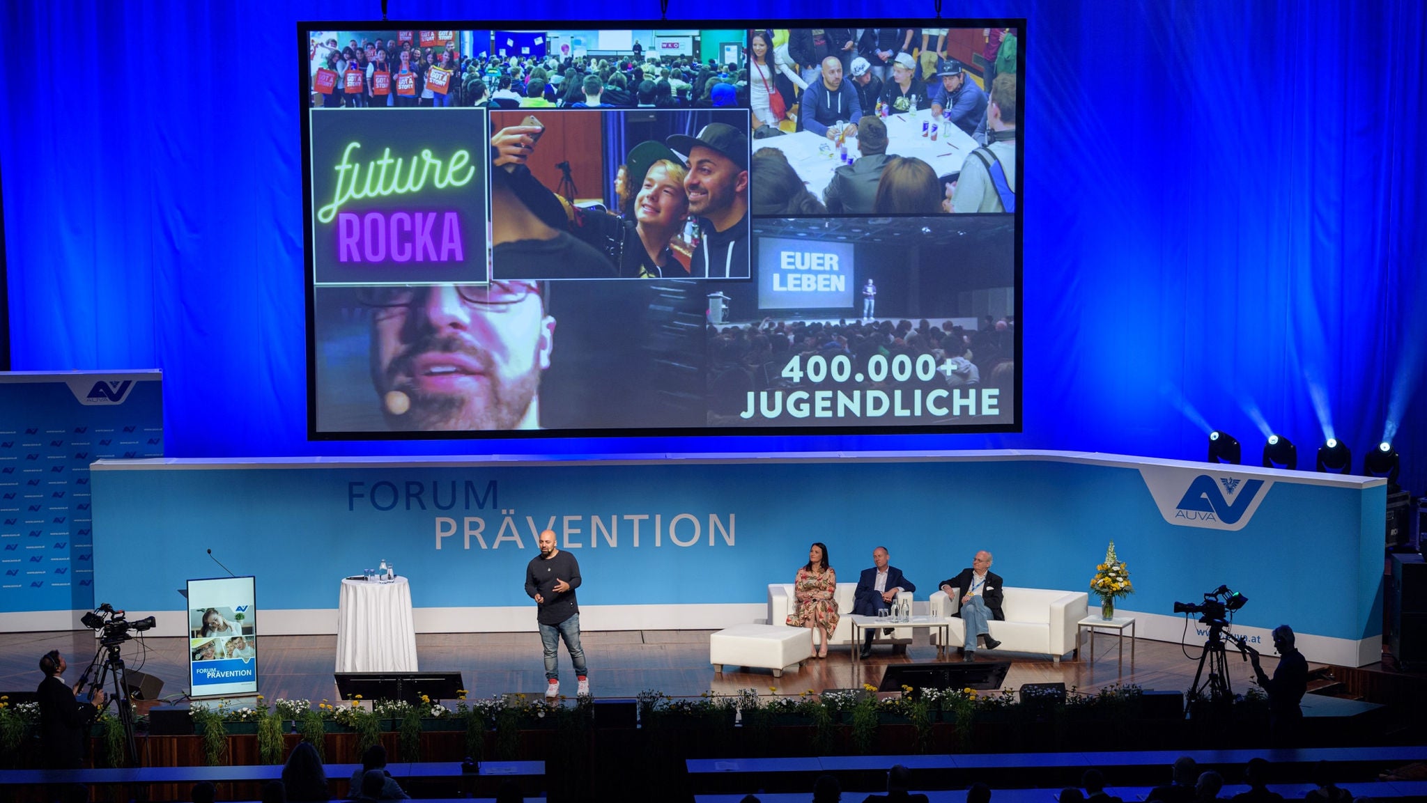Bühne mit Keynote-Speaker Ali Mahlodji, darüber ein großer Screen mit verschiedenen Einblicke in das Forum: Arbeitskreise mit Jugendlichen, Jugendliche, die Selfies machen, Vortragende mit Mikrofon, verschiedene Screens