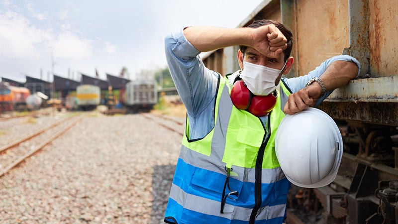 Eine Person in Warnweste und Mundschutz lehnt vor Hitze erschöpft an einem Zugwaggon und hält einen weißen Schutzhelm in der Hand.