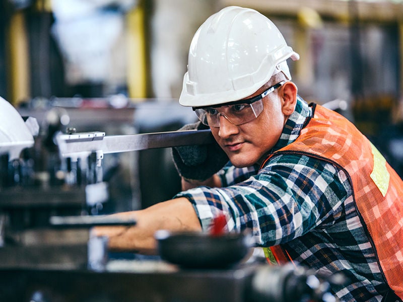 Eine Person mit Schutzhelm, Schutzbrille und orangefarbener Sicherheitsweste arbeitet konzentriert an einer Maschine in einer Werkstatt.