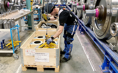 Eine Person in Arbeitskleidung und Schutzbrille bedient in einer Werkstatt eine Hebevorrichtung, um ein schweres Maschinenteil aus einer Holzkiste zu heben.