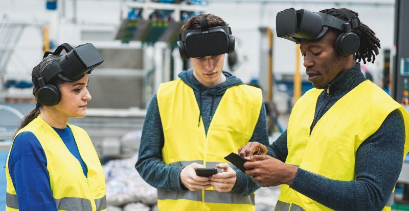 Drei Personen tragen Virtual-Reality-Headsets und gelbe Warnwesten in einer industriellen Umgebung. Zwei von ihnen halten Smartphones in der Hand, während die dritte Person aufmerksam zuhört. Die Szene deutet auf eine Schulung oder ein Training mit VR-Technologie hin, möglicherweise zur Simulation von Arbeitsprozessen oder zur Sicherheitsschulung in der Produktion. Im Hintergrund sind Maschinen und Produktionsanlagen erkennbar.