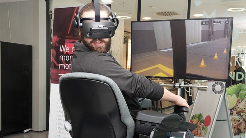 Virtuelle Trainingssituation für einen Gabelstaplerfahrer, der Mann mit virtuellem Helm dreht den Kopf und übt das Reversieren; davor zwei Monitore