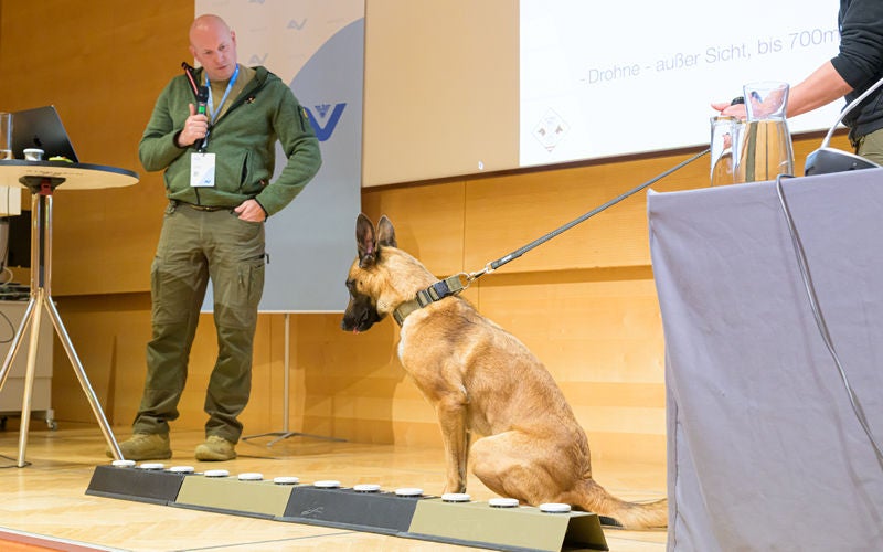 Mann mit Mikrofon hält einen Vortrag auf der Bühne neben einem Diensthund.
