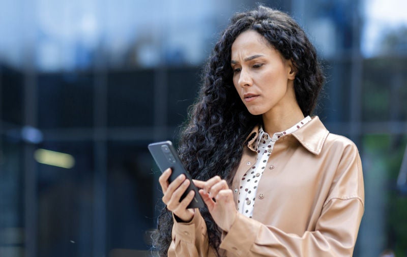 Eine Frau mit langen, lockigen Haaren und beigefarbener Jacke betrachtet konzentriert ihr Smartphone. Im Hintergrund unscharf gehalten eine urbane Umgebung. Ein Text beschreibt die potenziellen Auswirkungen mangelnder Transparenz bei digitalen Prozessen auf Unsicherheit, Misstrauen und Überwachungsgefühl.
