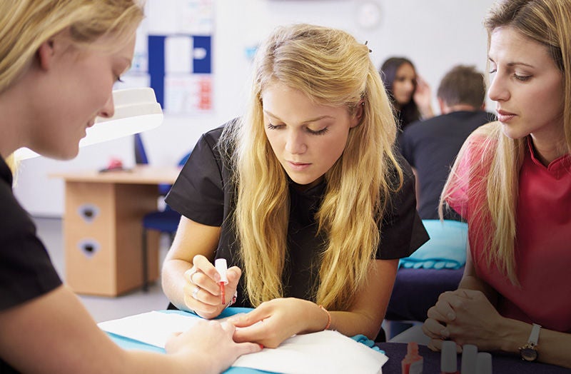 eine junge Nageldesignerin lackiert den Nagel einer Frau, eine andere Frau blickt ihr freundlich prüfend über die Schulter