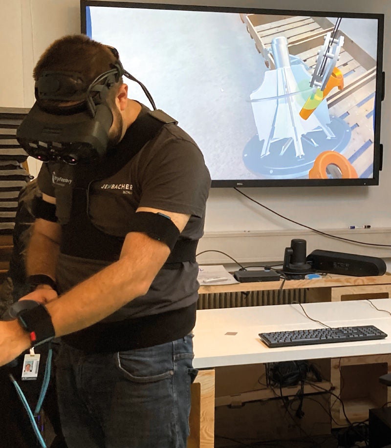 Person mit VR-Headset hebt virtuell ein Motorenteil mit einem virtuellen Kran und Lastaufnahmemittel, während auf einem Monitor die Simulation des Vorgangs dargestellt wird.