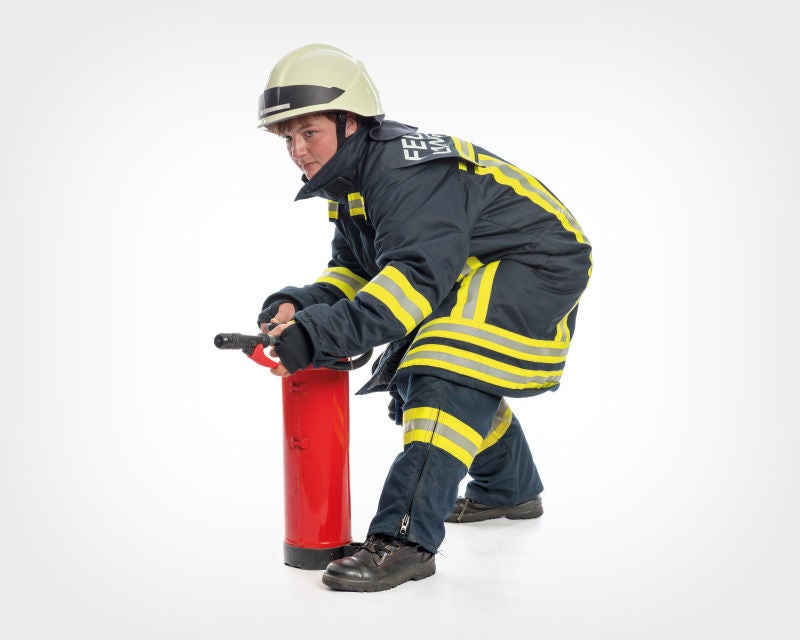 Ein Feuerwehrmann in voller Schutzkleidung, inklusive Helm, kniet nieder und hält einen Feuerlöscher, bereit, diesen zu benutzen. Der Hintergrund ist weiß, wodurch der Fokus vollständig auf den Feuerwehrmann und den Feuerlöscher gelenkt wird.