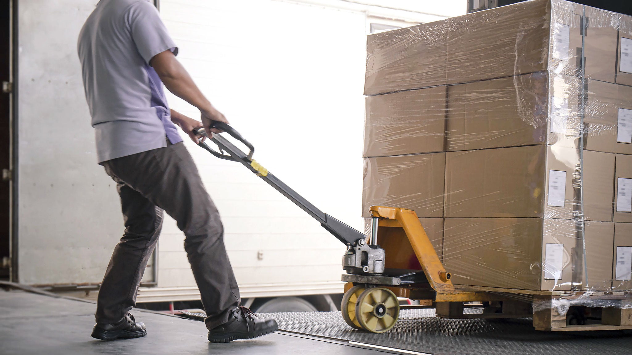 Ein Arbeiter müht sich, Lasten auf einer Palette mit Hilfe eines Handhubwagens auf dem Laderaum eines LKW auf die Laderampe zu verfahren