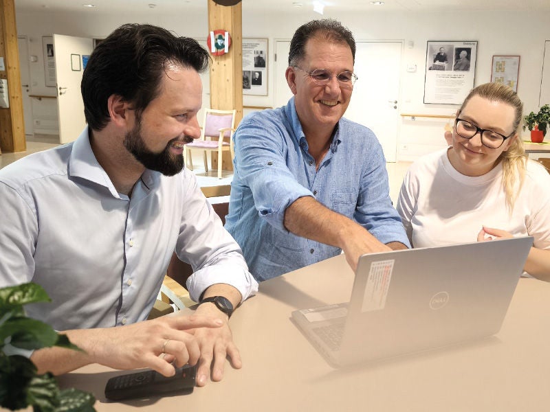 Zwei Männer und eine Frau sitzen an einem Tisch, schauen auf einen Laptop und diskutieren lächelnd in einer freundlich gestalteten Umgebung.