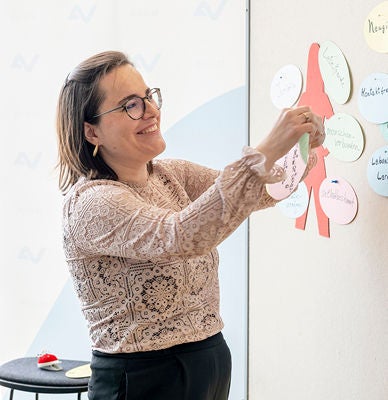 Eine Vortragende mit Brille und langem Haar steckt bunte Zettel an eine Pinnwand in einem hellen Raum.