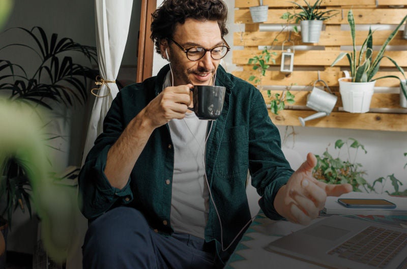 Ein Mann mit Brille und Kopfhörern hält eine Tasse in der Hand und lächelt. Er sitzt in einem Raum mit vielen Pflanzen, darunter einige in hängenden Töpfen und auf einem vertikalen Holzregal arrangiert. Ein Vorhang und ein Fensterrahmen sind im Hintergrund sichtbar.