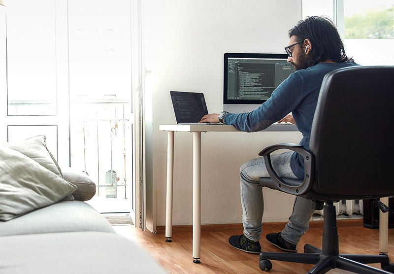 ein Mann sitzt an einem Schreibtisch mit Notebook und einem großen Monitor, rechts davon ein Sofa und im Hintergrund ein kleiner Balkon