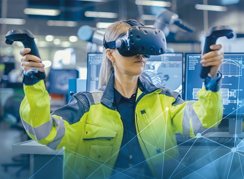 eine Frau in Arbeitskleidung und Schutzjacke mit einer VR Brille und zwei Sticks in den Händen, dahinter viele Monitore