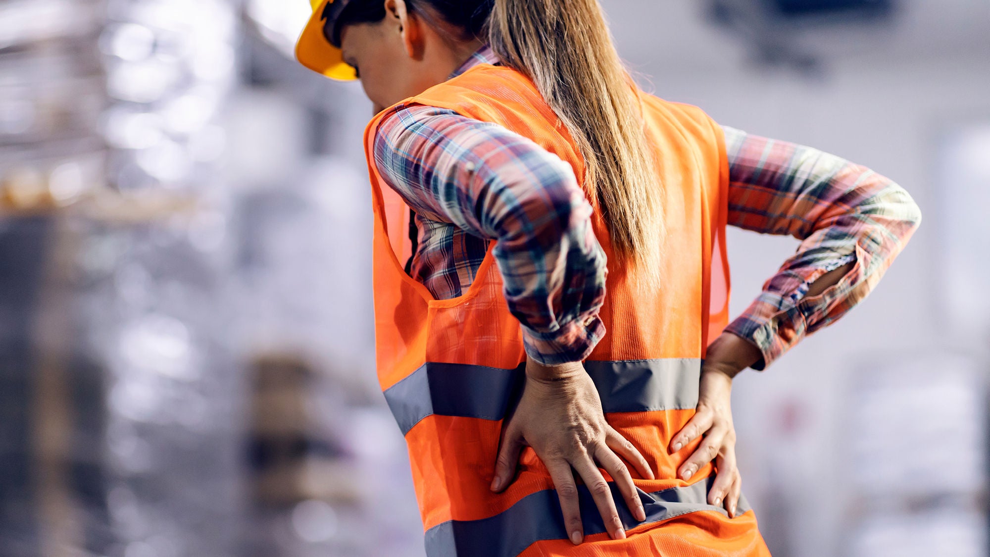 Eine Person mit langem Haar trägt eine orange Warnweste und einen Schutzhelm und hält sich den Rücken in einer Lagerhalle.